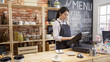 © PRPicturesProduction - smiling shop assistant using pos point of sale terminal to put in order from note at restaurant register. young girl waitress standing in wooden bar counter and working on digital tablet in cafe