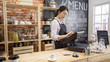 © PRPicturesProduction - small business people and service concept. young asian girl waitress at wood bar counter in cafe store. woman bartender in apron using pos sale terminal machine to enter customer order from note.