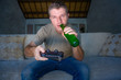 © TheVisualsYouNeed - indoors portrait of young excited and happy man at home playing videogames holding controller enjoying cheerful and drinking beer bottle in video gaming addiction