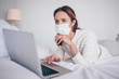© Алина Троева - Young european woman in face medicine mask working on a laptop in bedroom during coronavirus isolation home quarantine. Covid-19 pandemic Corona virus. Distance online work from home concept.