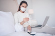 © Алина Троева - Sick woman in face protection mask lying in bed with laptop holding thermometer and pills at home quarantine isolation. Distance online work from home. Corona virus infection COVID-19 concept.