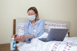 Close up shot of young blonde woman wearing disposable face mask working at home due to coronavirus quarantine concept. Beautiful female sitting in bed with laptop. Background, copy space.