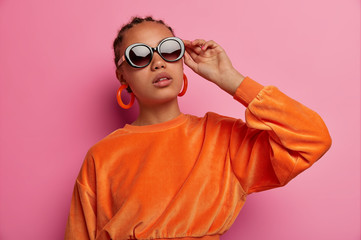 Wall Mural - Photo of confident dark skinned Afro American female model wears trendy sunglasses, dressed in orange sweater, prepares for date, isolated on pink background, has fashion look, cornrow hairstyle