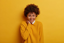 Beautiful Optimistic Woman With Smooth Skin And White Teeth Smiles Tenderly, Touches Neck, Raises Mood As Watches Comedy At Home, Wears Bright Yellow Jumper, Sends Good Vibes, Models In Studio