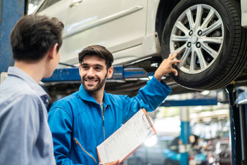 Vehicle maintenance handsome man servicer explain car condition, point at vehicle part to Asian customer in garage. Automotive mechanic hold automotive checklist document. Car repair service concept.