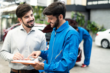 Wall Mural - Vehicle maintenance handsome man servicer explain car condition and expense to smile male customer in garage before sign automotive repair quatation document. Vehicle repair service business concept.