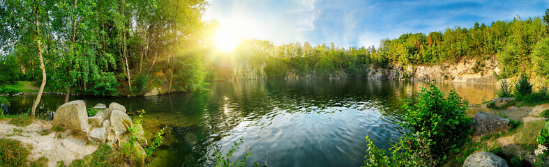 Wall Mural - Panoramic landscape shot of idyllic lake surrounded by trees and cliffs, with the sun glowing on the horizon