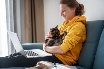 Happy young woman in a yellow sweatshirt works at home on a blue sofa with a laptop and a cat, remote work and education, staying home during a virus