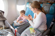 a little cute toddler girl sitting in an airplane in a chair by the porthole herself wipes a folding table with a wet antibacterial napkin before eating. mom applauds and praises the child