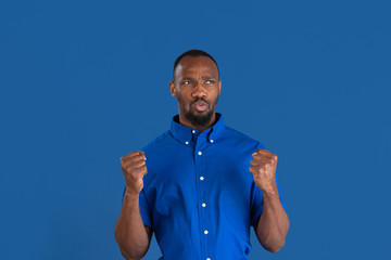 Wall Mural - Celebrating win. Monochrome portrait of young african-american man isolated on blue studio background. Beautiful male model. Human emotions, facial expression, sales, ad concept. Youth culture.