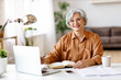 © JenkoAtaman - Senior woman with laptop and notebook smiling and looking at camera while sitting at table and working on remote project at home.