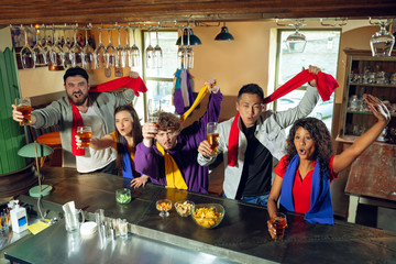 Wall Mural - Sport fans cheering at bar, pub and drinking beer while championship, competition is going. Multiethnic group of friends excited watching translation. Human emotions, expression, supporting concept.