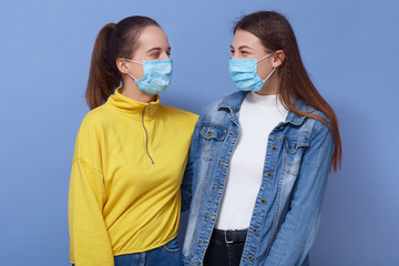 two omen wearing disposable protective face mask and casual outfits. Coronavirus, covid 19, virus epidemic concept, girls friends hugging and looking at each other, isolated over blue background.