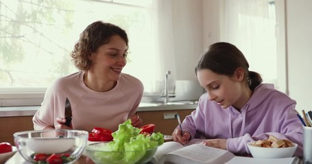 Wall Mural - Friendly family young mother and teenage daughter enjoying lifestyle at home. Adult parent mum cutting vegetables preparing salad while school girl child studying doing homework sit at kitchen table.