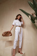 canvas print picture - Beautiful woman fashion model brunette hair tanned skin wear white overalls button suit sandals high heels accessory bag clothes style journey safari summer collection plant flowerpot wall stairs.