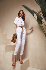 Wall Mural - Beautiful woman fashion model brunette hair tanned skin wear white overalls button suit sandals high heels accessory bag clothes style journey safari summer collection plant flowerpot wall stairs.