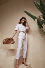 Wall Mural - Beautiful woman fashion model brunette hair tanned skin wear white overalls button suit sandals high heels accessory bag clothes style journey safari summer collection plant flowerpot wall stairs.