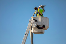 Man In Manlift Bucket Repairing Street Light Against Blue Sky