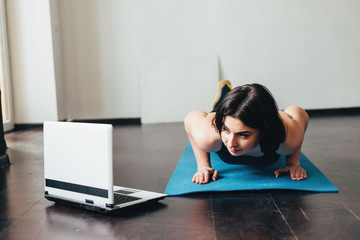 Home training,sport, online fitness class. Young fit woman doing exercise on mat looking video guide on laptop