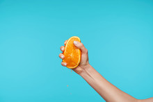 Colorful Shot Of Raised Female's Hand With Half Of Fresh Orange, Smashing It With Fingers While Making Juice, Posing Over Blue Background. Hands And Gesturing Concept