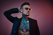 © Fxquadro - Bold and rebellious male model posing in a neon studio wearing a black tuxedo on a half-naked body, fire-shaped sunglasses and tattooed in a japanese irezumi style, looking self-assured.