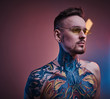 © Fxquadro - Self-assured male model posing in a neon studio with a half-naked body wearing yellow sunglasses and tattooed in a japanese irezumi style, looking cool and relaxed. Close up portrait