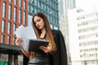 © bodnarphoto - Closeup portrait of pretty young businesswoman in dark formal wear studying business papers against a skyscraper background, concentrating on paper, holding a laptop in her hand. Business concept.