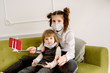 © irishasel - Two girls play a doctor at home in a bright room. On a green sofa they play with a bear, bandage a foot to a toy. In a face mask, protection against Chovid-19 virus.Big sister takes selfie for blog