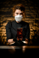 Wall Mural - Close-up. Woman at bar in medical mask and black gloves present glass with cocktail.