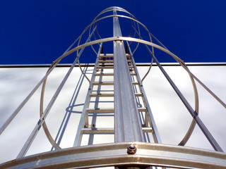 aluminum roof access ladder with safety cage in diminishing perspective. strong shadows on white concrete wall. rungs and vertical safety bars. buildings, architecture and technology. clear blue sky.