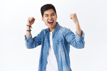 Wall Mural - Portrait of empowered, enthusiastic asian man fist pump, raise hands up in triumph, celebrating victory, smiling saying yes, winning game or competition, achieve goal, white background