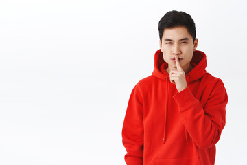 Wall Mural - Portrait of devious good-looking asian man in red hoodie, keep secret, squinting mysterious and shush, make hush gesture, finger to lips, hiding something, prepare surprise, white background