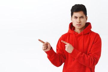 Wall Mural - Portrait of young asian man looking with judgement and disappointment at camera, frowning angry pointing fingers left, demand answers, being displeased and outraged with something, white background