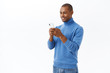 © Cookie Studio - Technology, online lifestyle concept. Portrait of handsome african-american man using mobile phone, texting friend, scrolling internet social network, smiling at smartphone display, white background