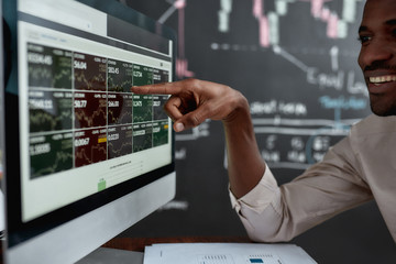 Wall Mural - Utilising skills for earnings. African businessman, trader sitting in front of computer screen and looking at graph chart with a smile. Blackboard full of data analyses in the background.