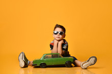 Kid In Sunglasses, Camouflage T-shirt, Sneakers. Sitting On Floor, Holding Green Model Of Retro Car, Posing On Orange Background