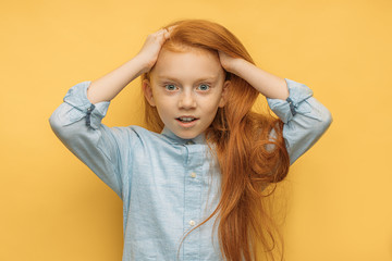 caucasian surprised red haired girl isolated over yellow background. adorable child with big blue eyes surprisingly react on something, girl grabbed hair and look at camera. positive emotional child