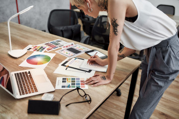 Wall Mural - Creative female. Young blonde tattooed female designer making some sketches while standing near office desk. Working with color swatch samples