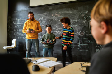 A Quality School. Young male science teacher standing near the blackboard, holding tablet pc and looking at his students while they are demonstrating their own robot vehicle