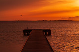 Fototapeta Pomosty - Wooden pier on a warm light during sunset, with orange sky and a Kite surf and two mountains silhouette in the background
