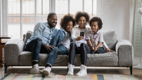 Horizontal image happy African ethnicity young family with small children having fun using smartphone laughing watching funny video on-line, talk distantly by video call, making selfie picture concept