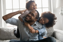 Happy African Family At Home, Cute Son And Daughter Hanging On Daddy Back, Father Fool Around With Little Son And Daughter Piggy Back Siblings Enjoy Active Time Together Seated On Sofa In Living Room