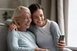 © fizkes - Happy millennial girl and senior mom sit on couch at home smile for self-portrait picture on cellphone together, overjoyed mature mother and adult daughter relax on sofa take selfie on smartphone