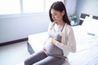 © Have a nice day  - beautiful asian pregnant woman smiling and hands rubbing firmly on her baby stomach, joyfully and happiness, sitting on the white bed relaxing and resting in the bedroom with light shining from window