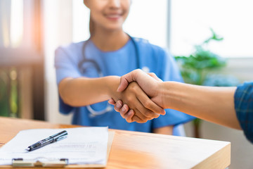 Wall Mural - Close up of asian female doctor shaking hands with patient in greeting and respect of doctor helping and support, with document forms and contracts or patients on desk within a hospital office room