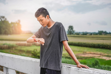 Wall Mural - male asian teenager looking at watch checking running distance and heart rate after running and exercising, wearing shirt, short and running shoes, with tree, nature and cloudy sky in the background