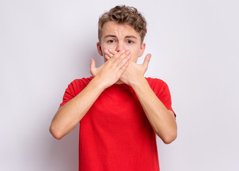 Wall Mural - Portrait of amazed teen boy covers his mouth with hands. Child in surprise looking at camera. Caucasian young teenager, on grey background. Speak no evil concept.