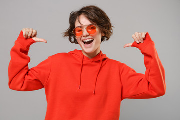 Funny young brunette woman girl in casual red hoodie, eyeglasses posing isolated on grey background studio portrait. People lifestyle concept. Mock up copy space. Pointing thumbs on herself, blinking.