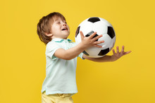 Funny Little Kid In Casual Summer Outfit Try To Catch The Ball Soccer Ball Over Yellow Background.