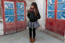 Young Woman On A City Street Dressed In A Black Coat, Flower Skirt, And Sunglasses, In Front Of Two Red Doors With Windows Covered In Broken Blue Plastics.